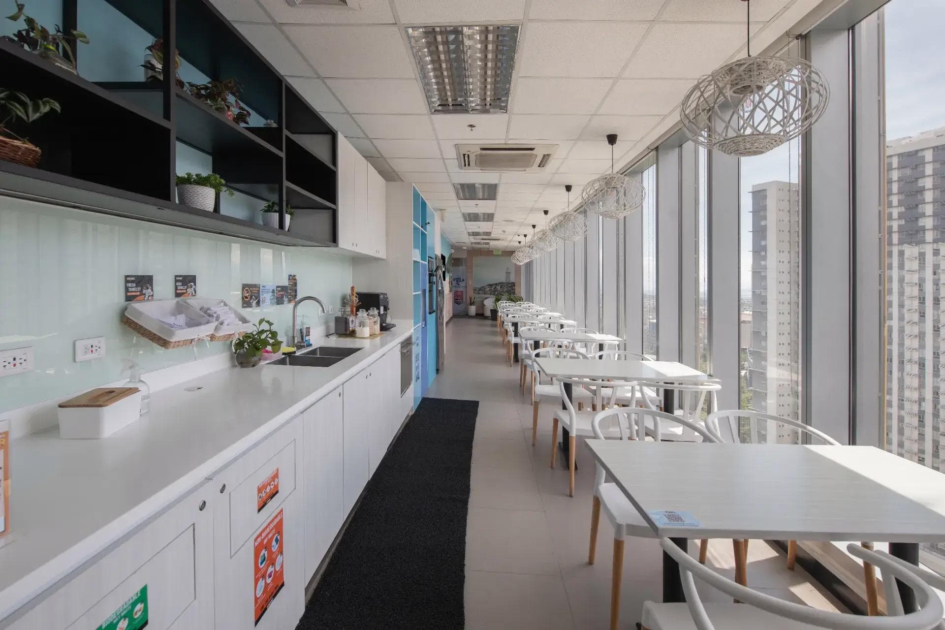Bright KMC pantry with white tables, long counter, and city views through tall glass windows in a BGC office.