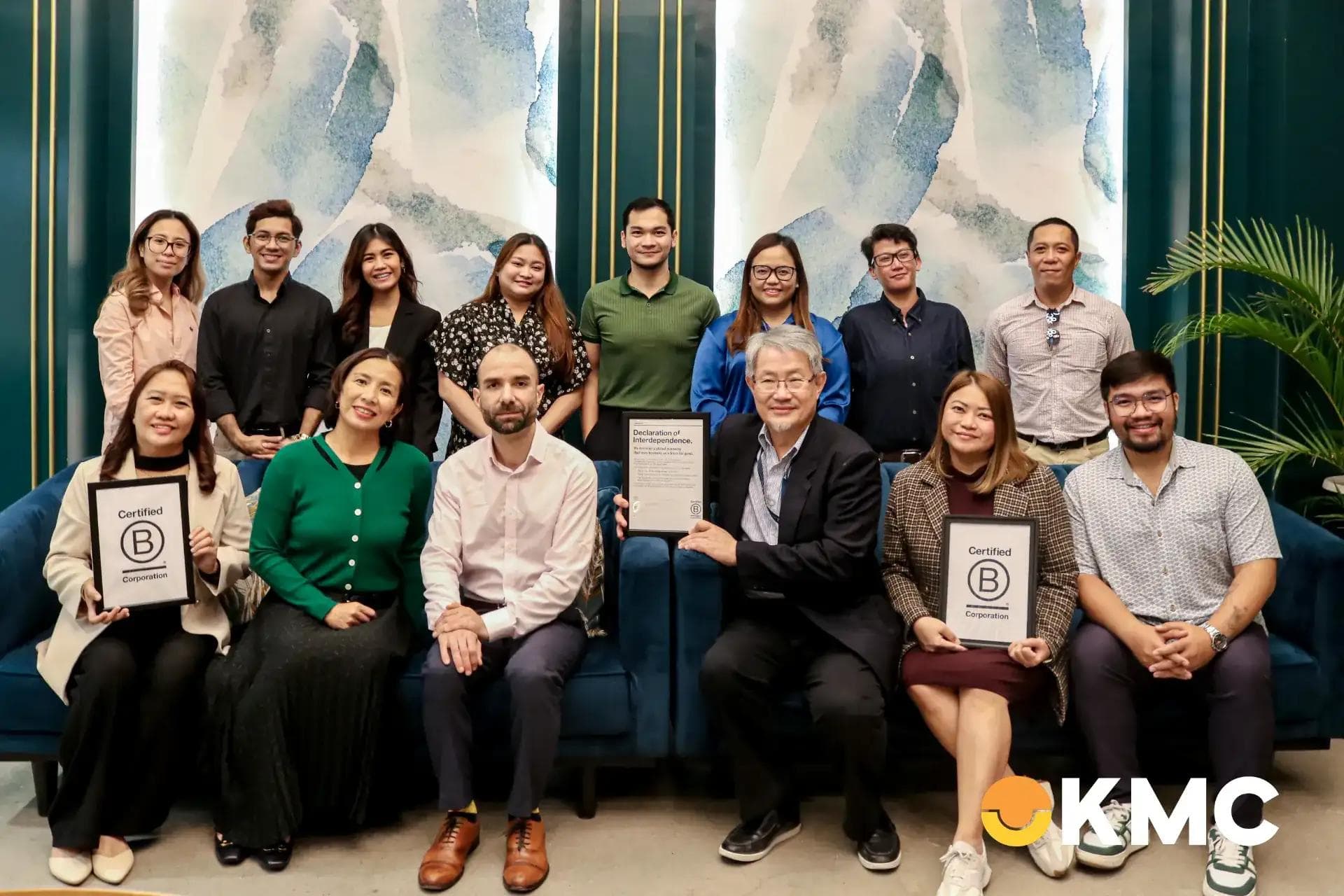 Group photo of KMC team members holding B Corp certification plaques, posing in front of a modern blue-green lounge backdrop.