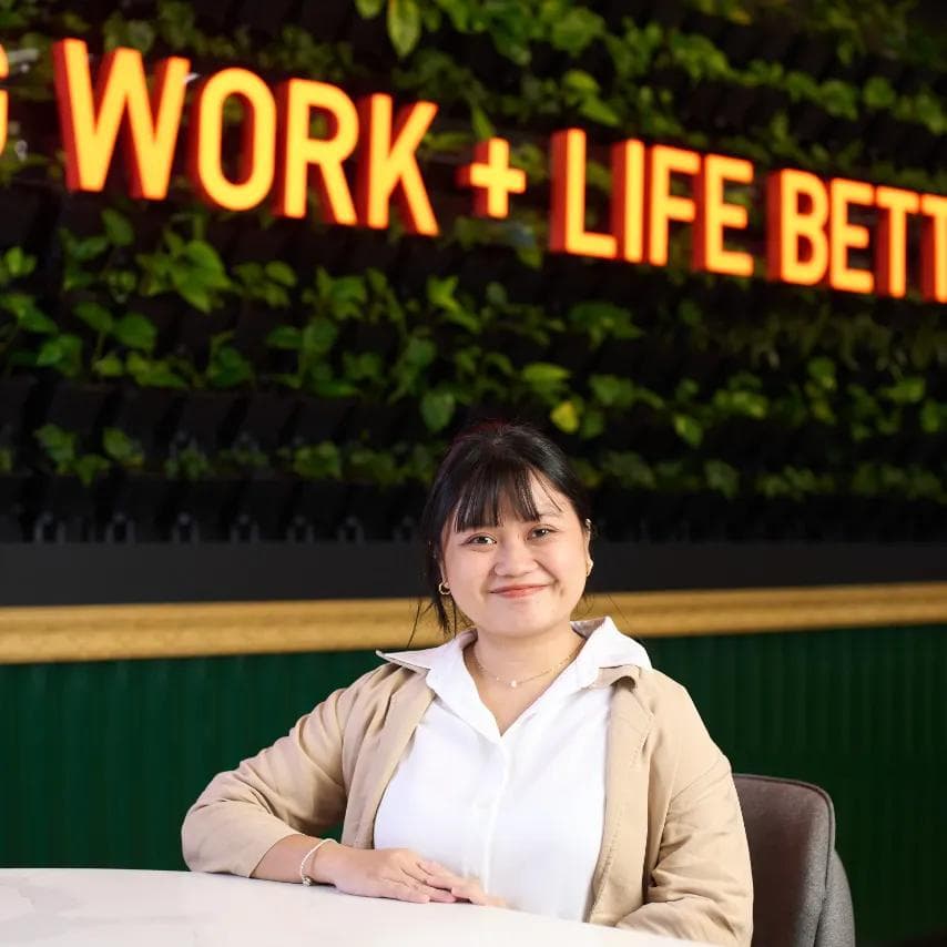 Woman seated at a round table in front of a lush green wall with a neon “Work + Life Better” sign, in a KMC office lounge.