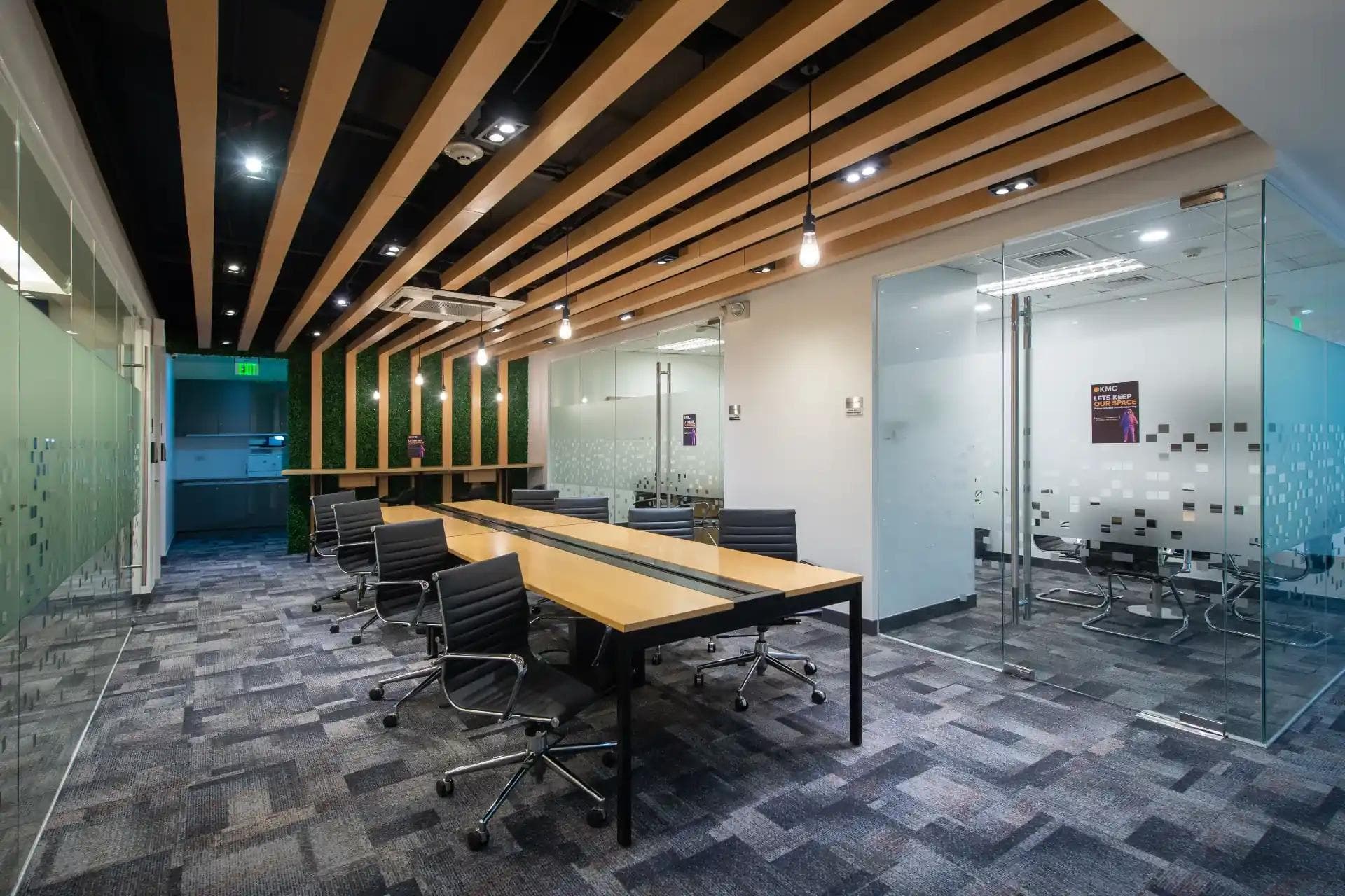 Modern office space with long wooden desks, black chairs, and glass-walled meeting rooms under wood-slat ceiling design.