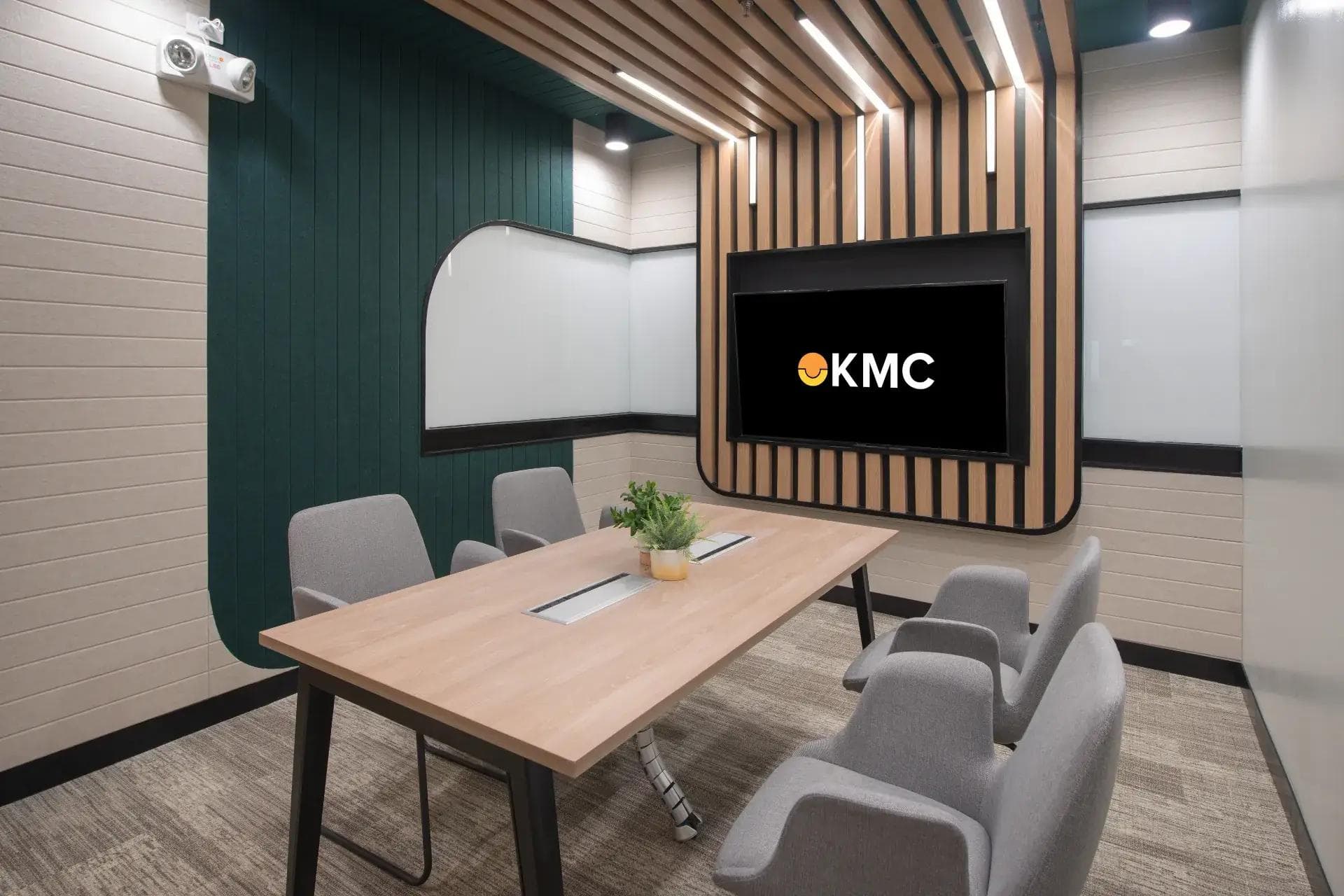 A stylish conference room with wood-accented ceiling, modern lighting, a large KMC-branded screen, and seating for four.