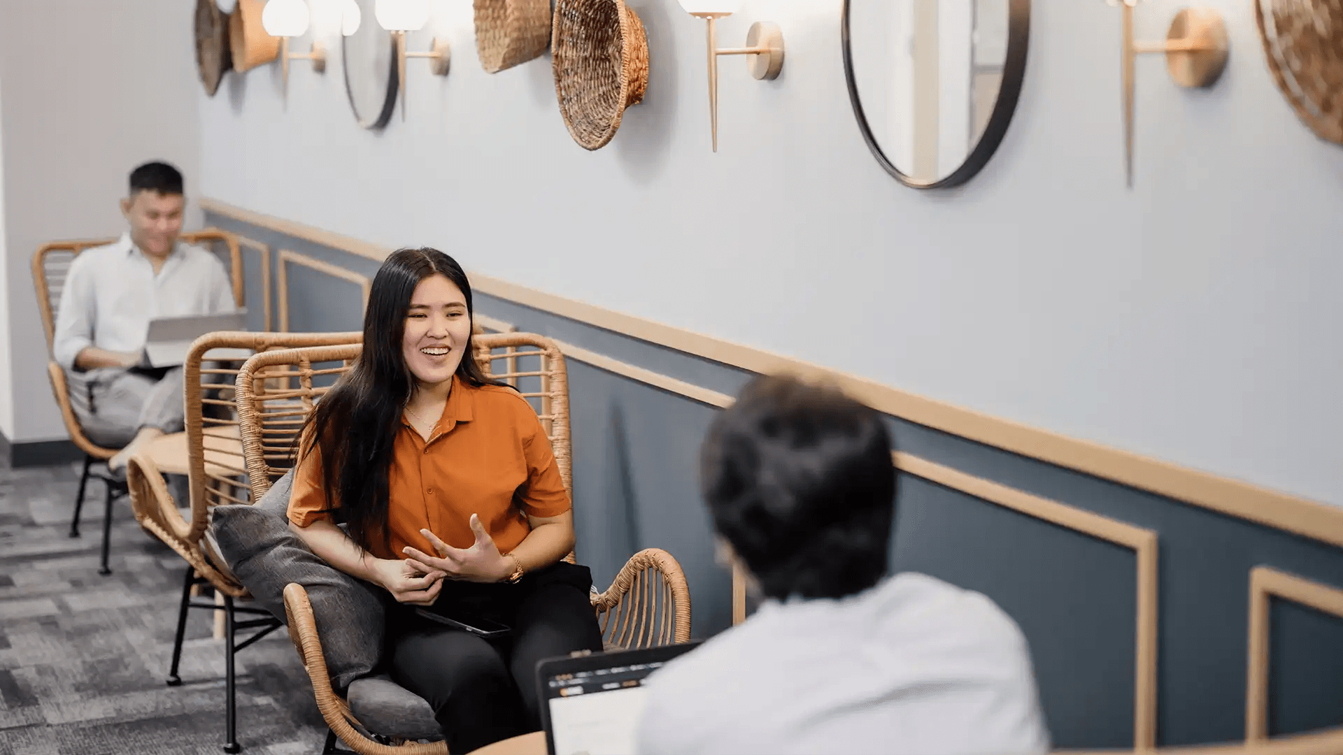 Professionals chatting in a cozy lounge with rattan chairs, round mirrors, woven wall decor, and a relaxed, modern vibe.