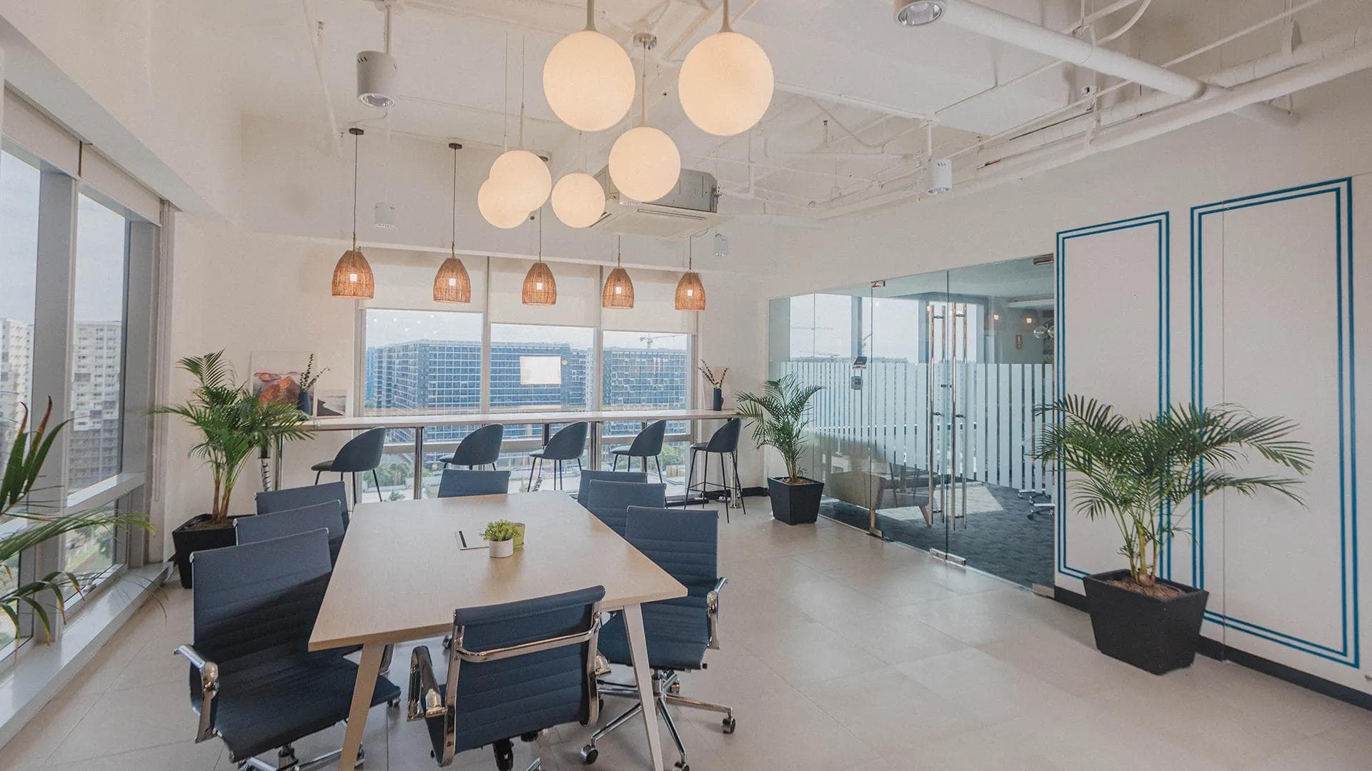 Modern meeting room with natural lighting, a long wooden table, indoor plants, and pendant lights in a KMC office space.