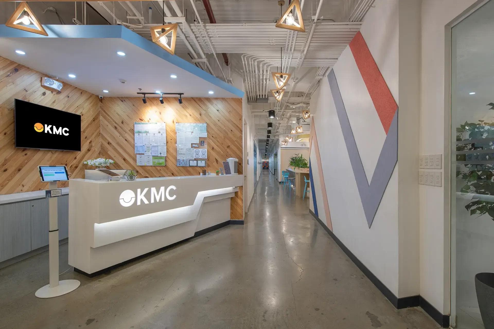 Office space in Cebu with geometric lights, wood accent wall, illuminated KMC logo, modern furnishings, and a sleek hallway.