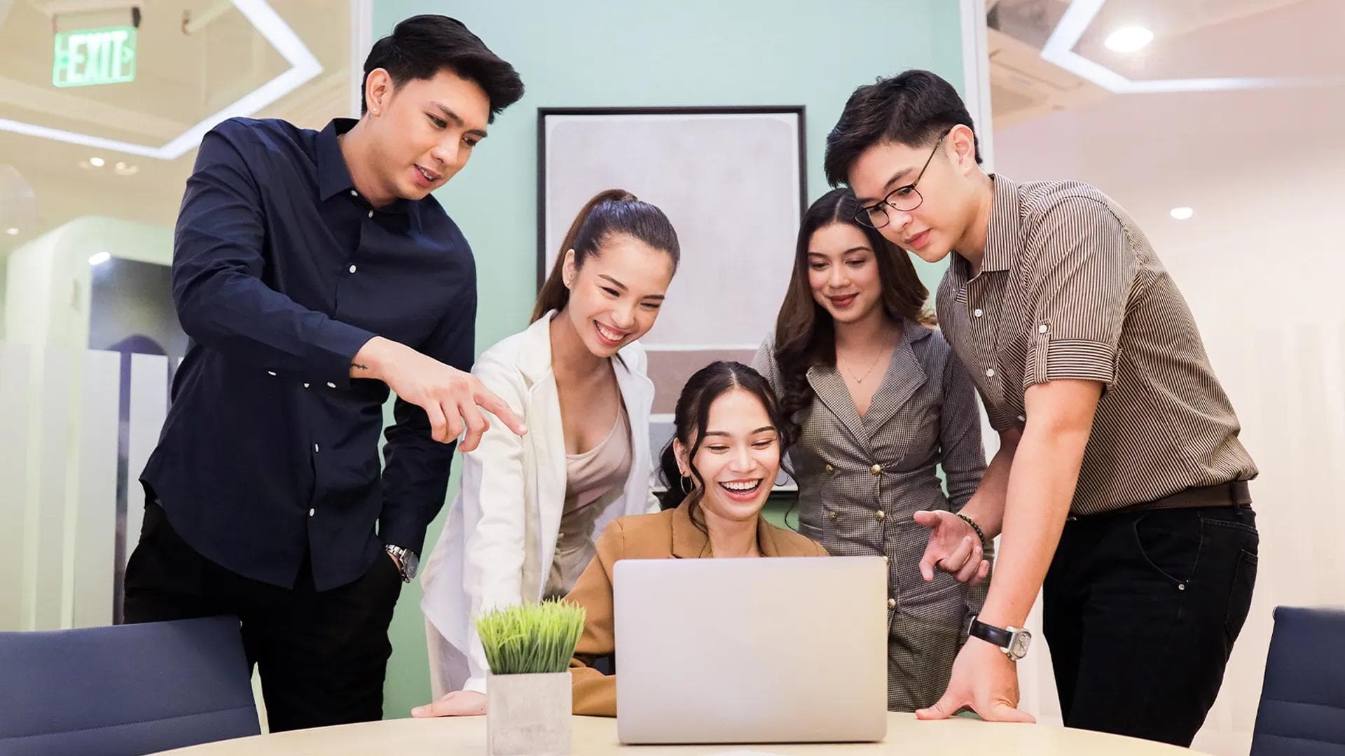 Five young professionals collaborating around a laptop in a modern office, representing outsourcing in the Philippines.