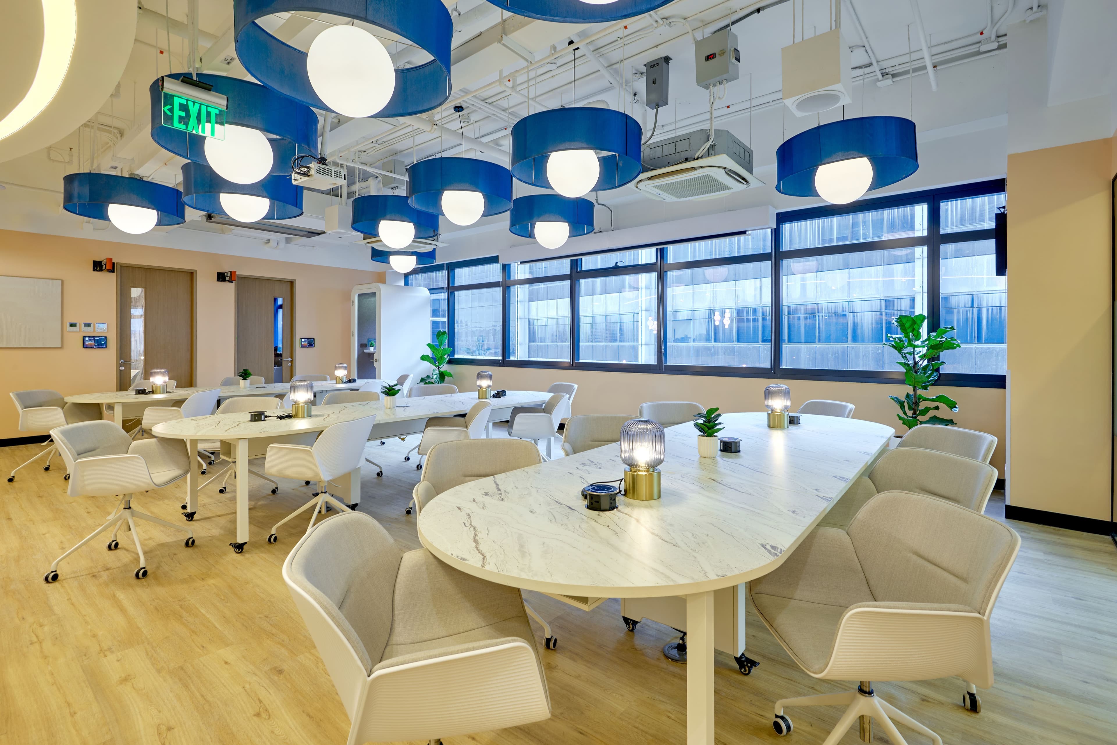 Bright coworking space with long white tables, modern chairs, blue ceiling lights, large windows, and lush green plants.