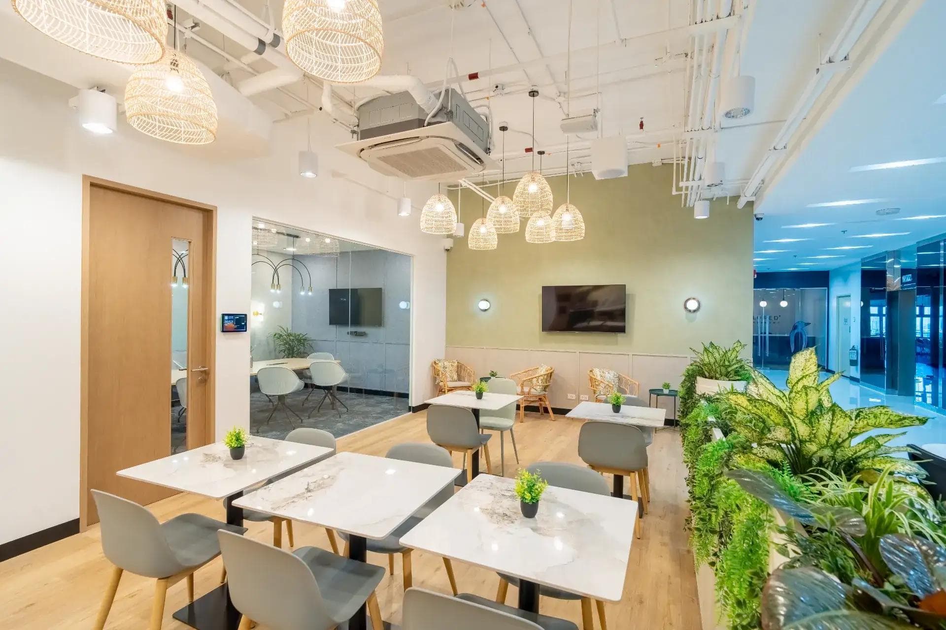 Bright coworking lounge with marble tables, six gray chairs, pendant lights, greenery, wood floors and a glass meeting room.