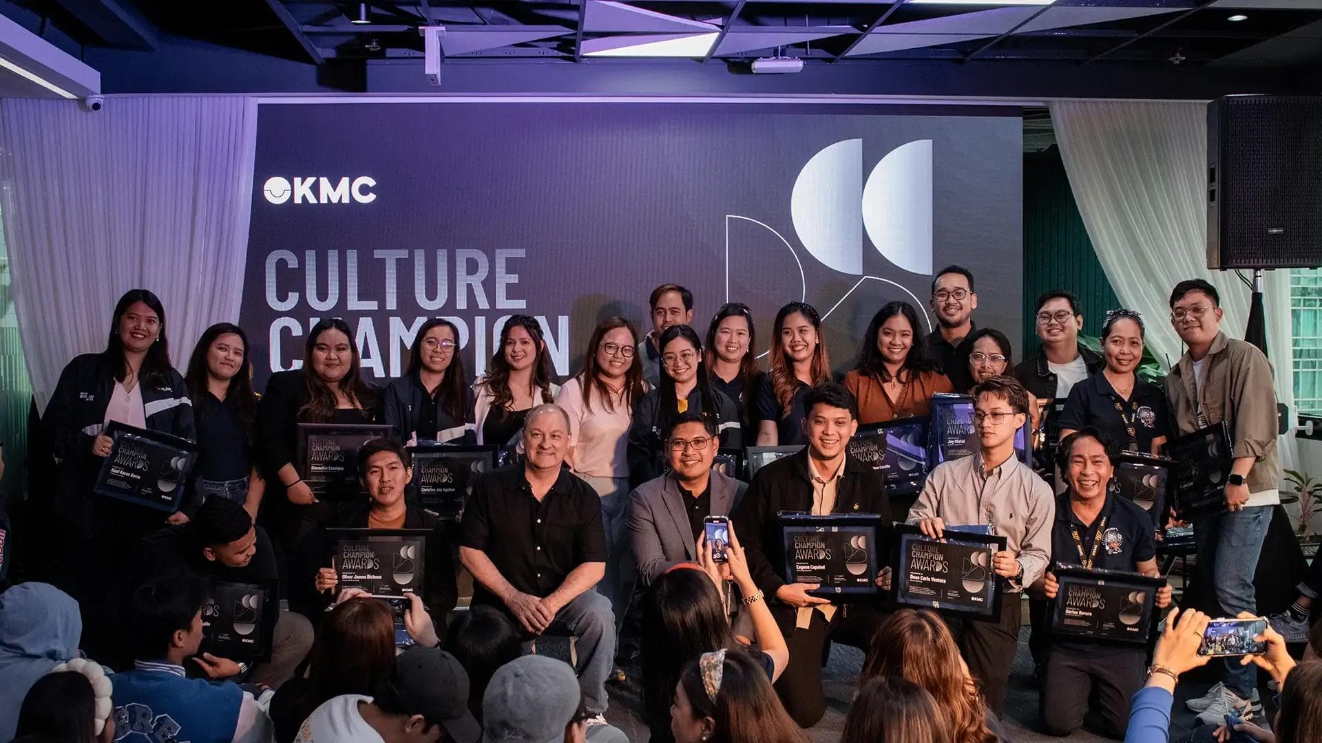 KMC team members celebrate Culture Champions awards on stage, smiling and holding plaques during an office event.