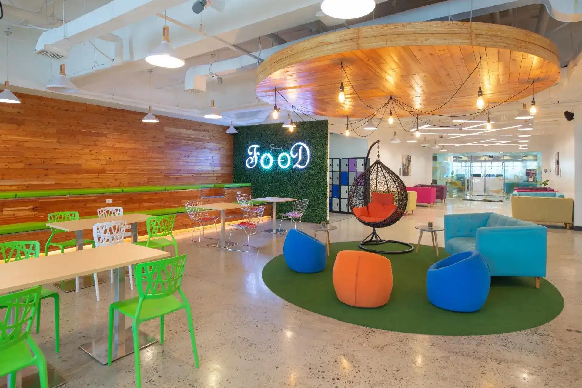 Colorful cafeteria lounge with modern seating, neon "FOOD" sign, hanging chair under a circular wooden ceiling feature.