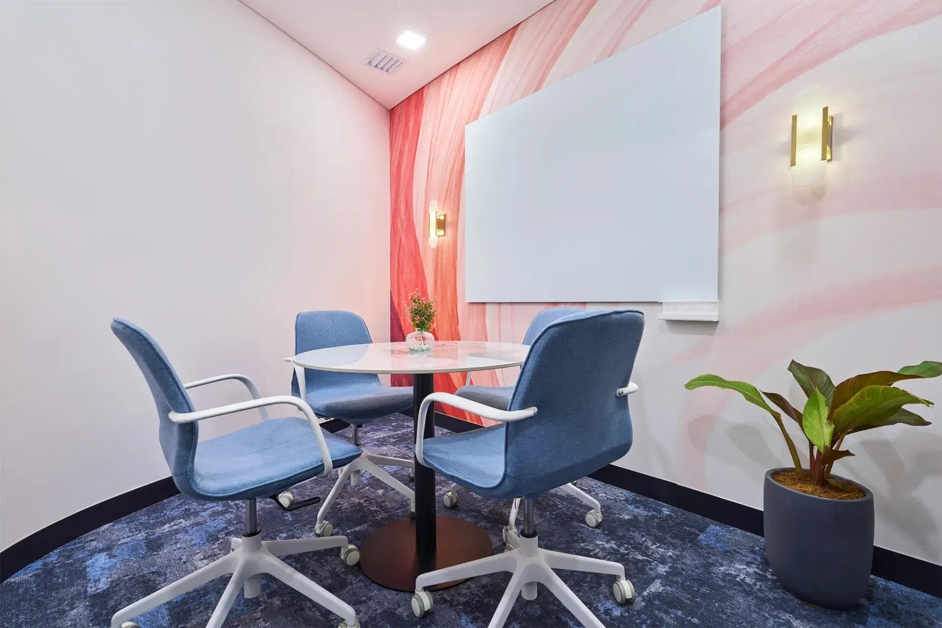 Private meeting nook with three blue chairs, round table, pink wall mural, whiteboard, potted plant, and brass sconces.