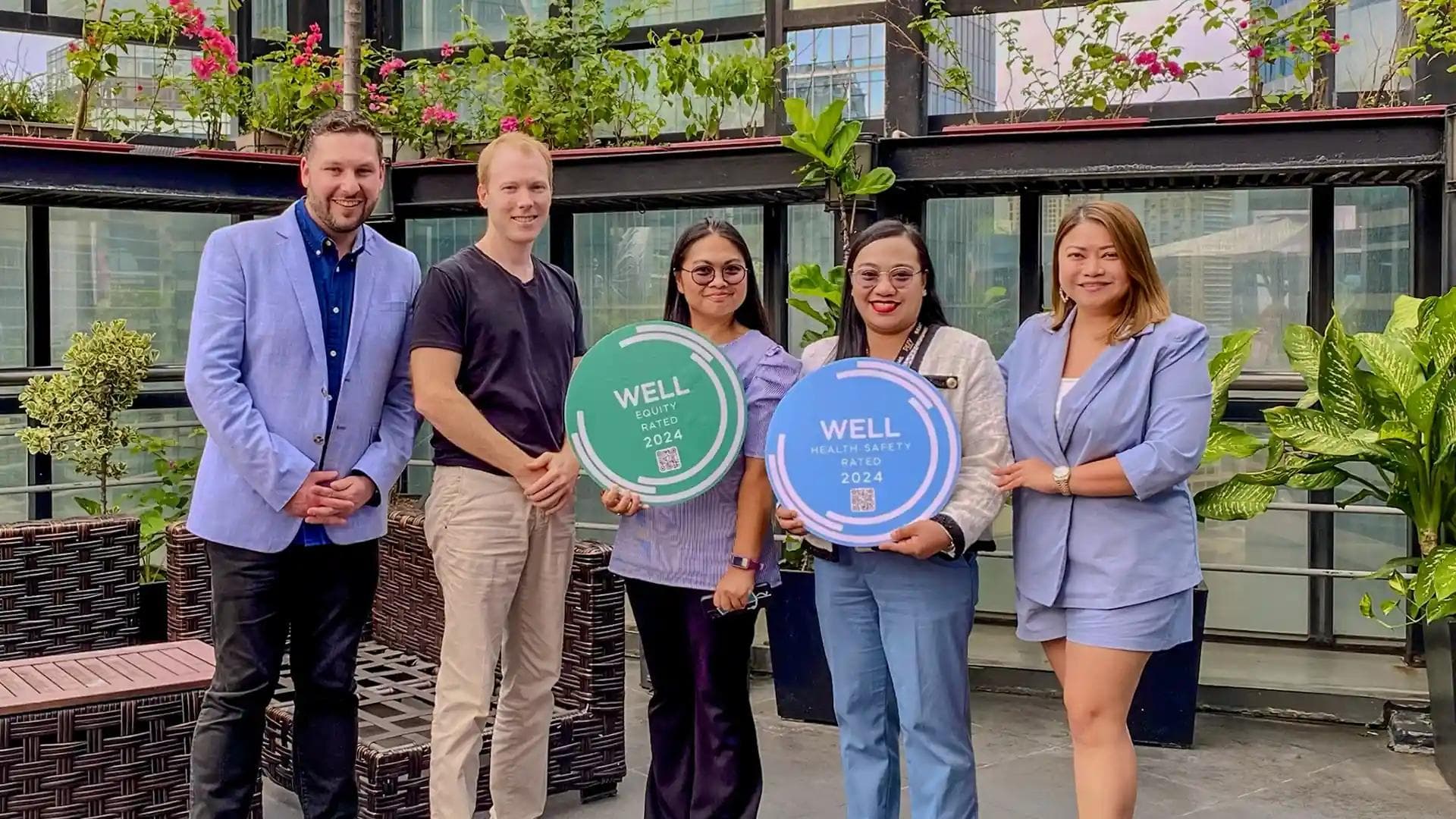 KMC team proudly holds WELL Certification seals while posing together on a rooftop with greenery in the background.