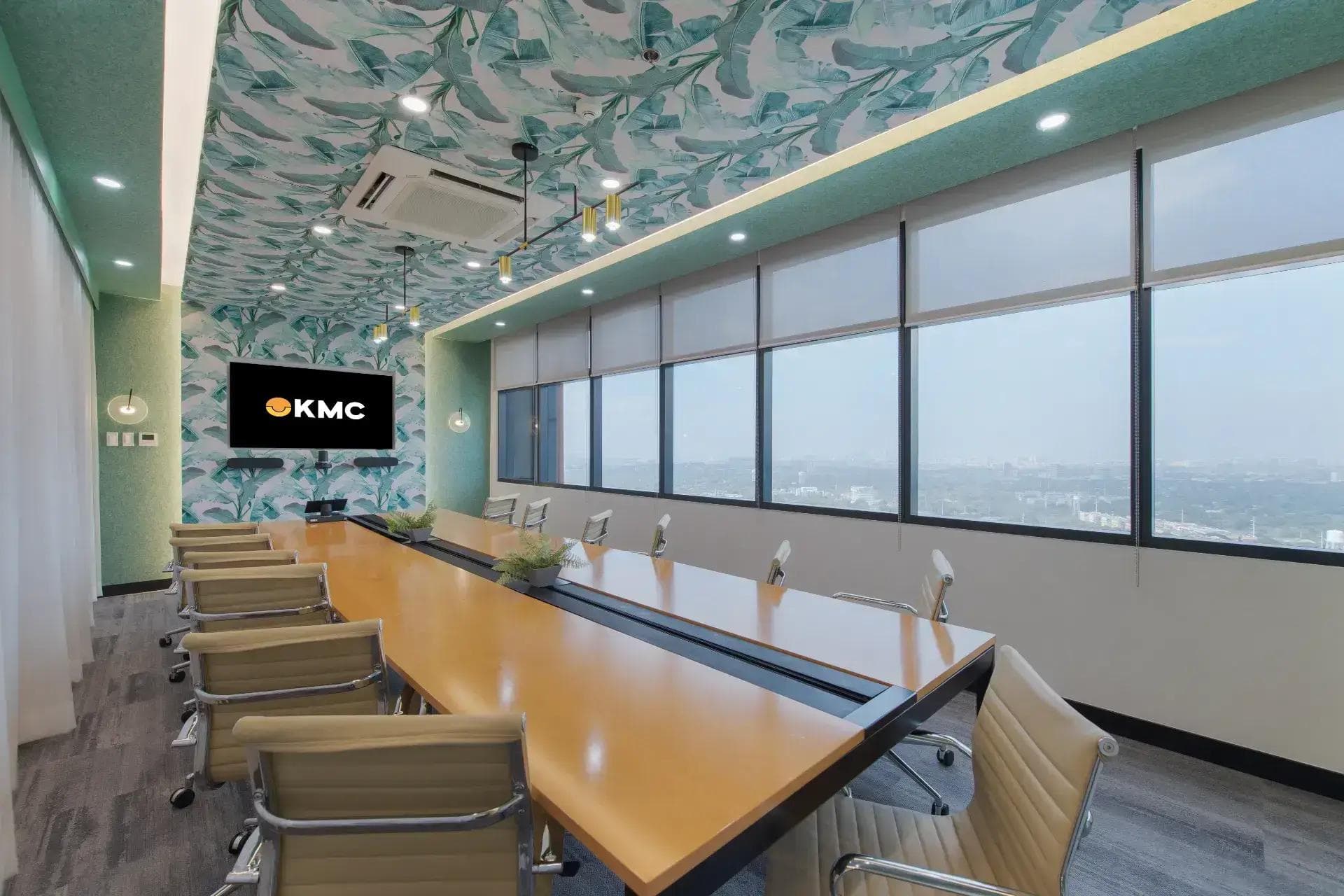 Bright KMC boardroom: long wood table, six gray swivel chairs, wall screen, wide windows, and a teal leaf-patterned ceiling.