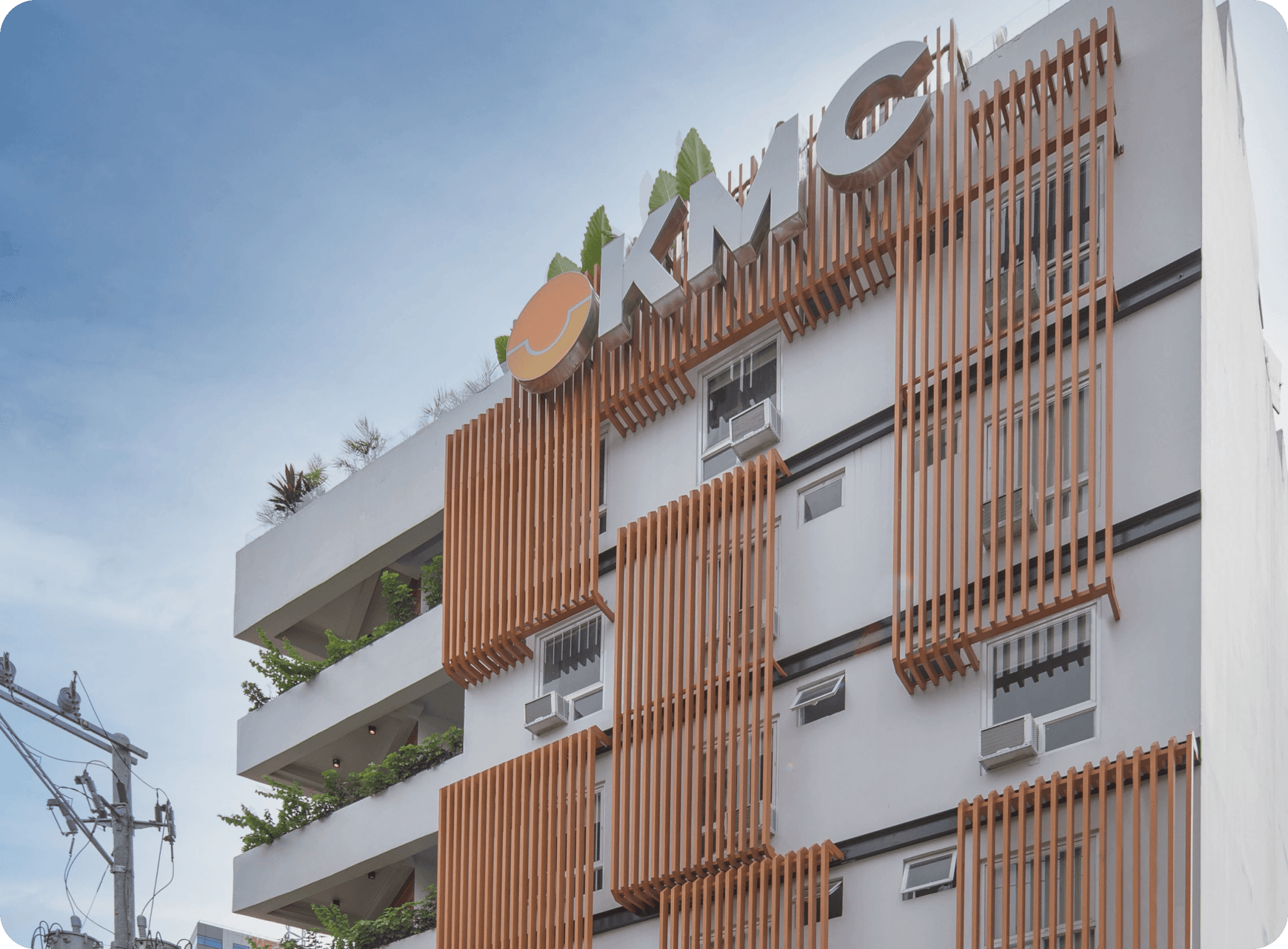 Exterior of Casa KMC with vertical wooden slats, modern design, greenery on balconies, and the KMC logo on the rooftop. Exterior of Casa KMC with vertical wooden slats, modern design, greenery on balconies, and the KMC logo on the rooftop.