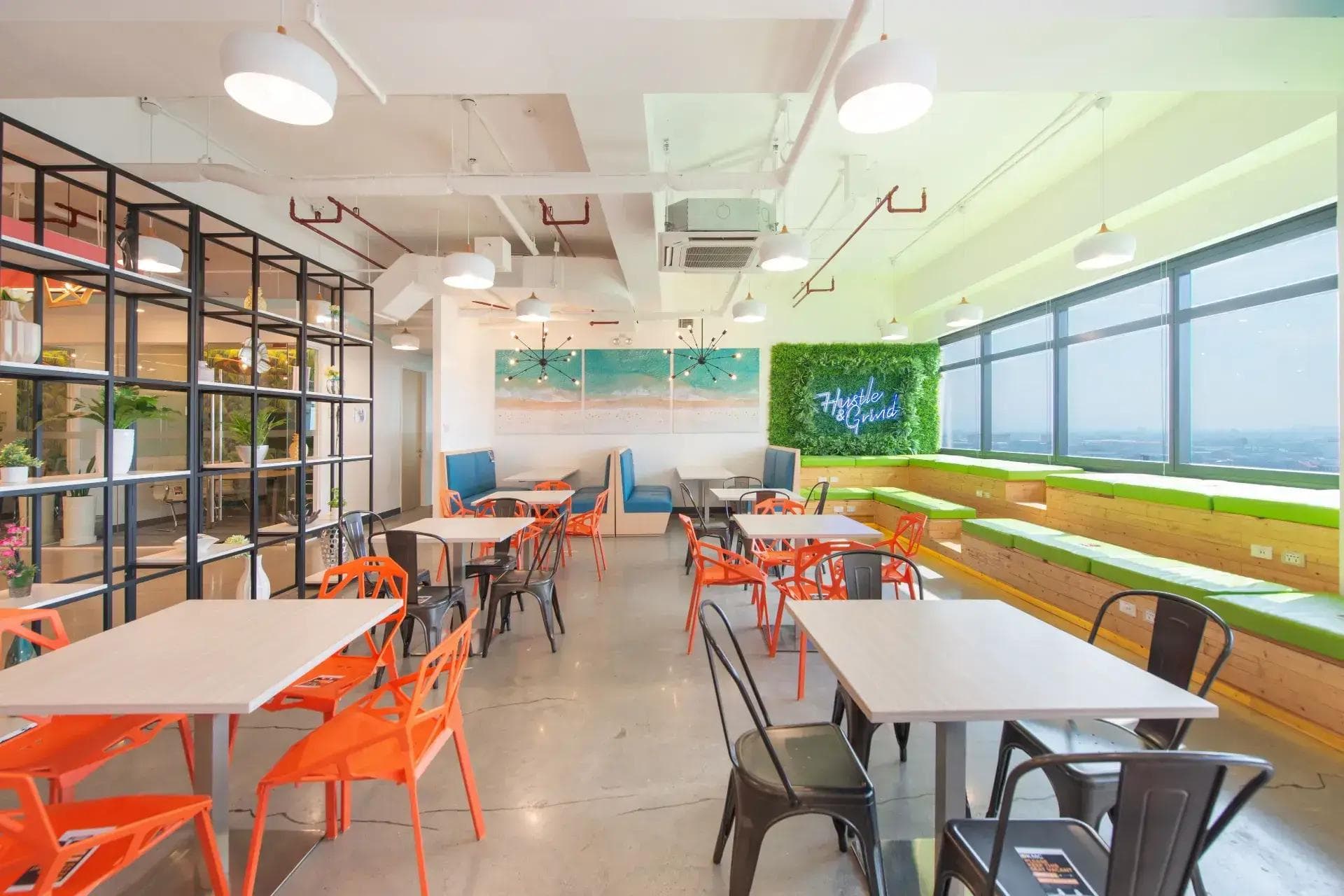 KMC’s coworking space with four light wood tables, orange and black chairs, booth seating, greenery, and large windows.