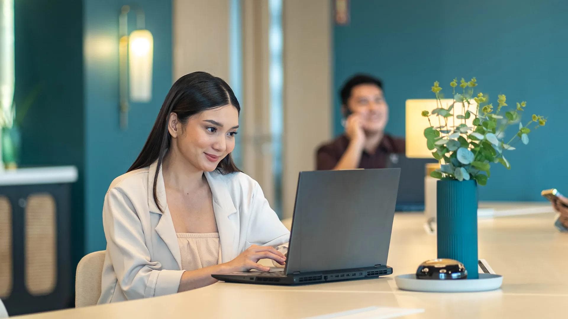 Smiling Filipino woman working on a laptop in a modern office lounge with lush decor and colleagues in the background.